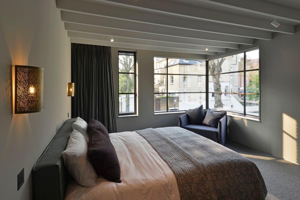 Modern bedroom with large windows, grey bed, and cozy seating area.