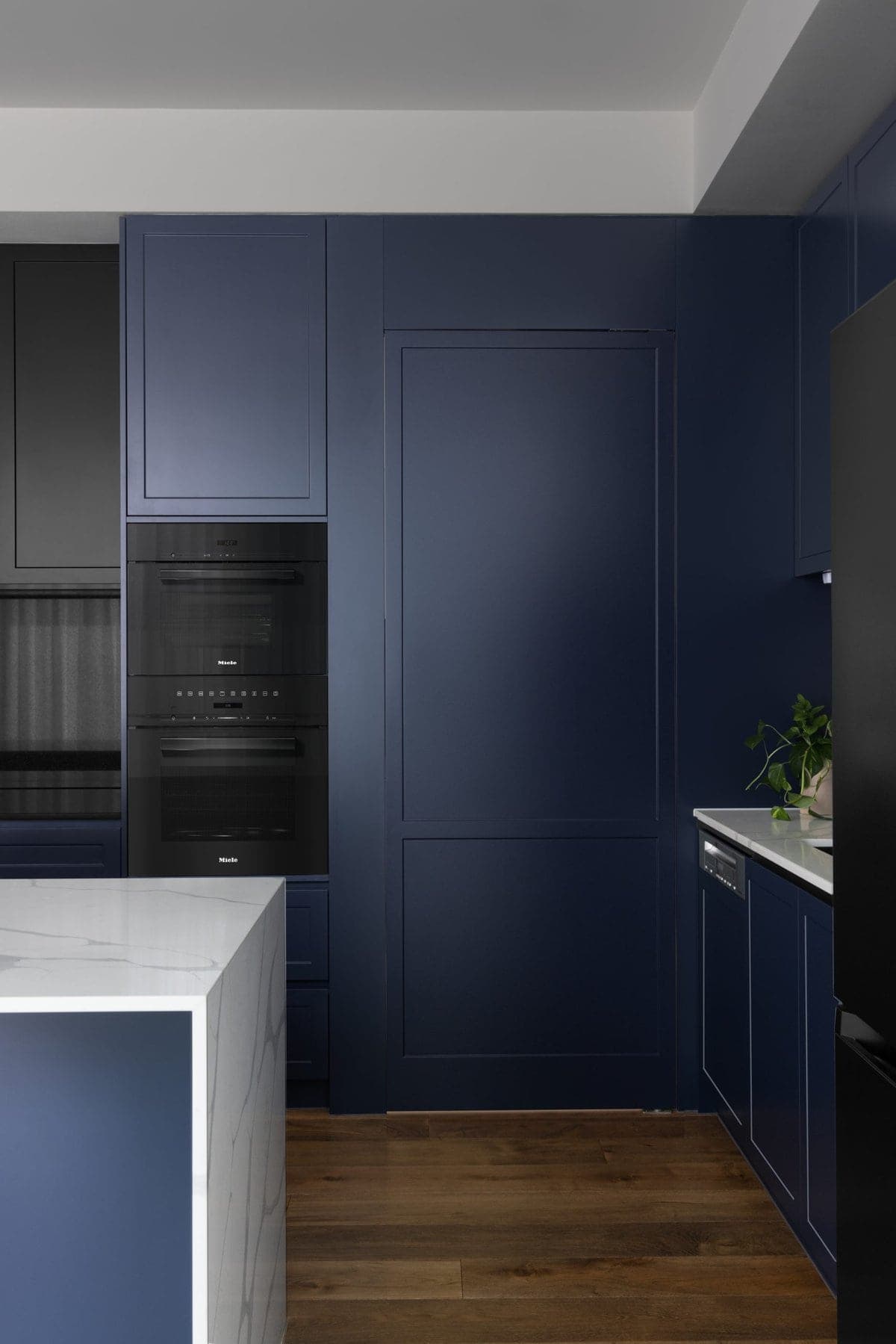 Sleek modern kitchen with dark blue cabinetry, white marble island, and built-in black appliances.