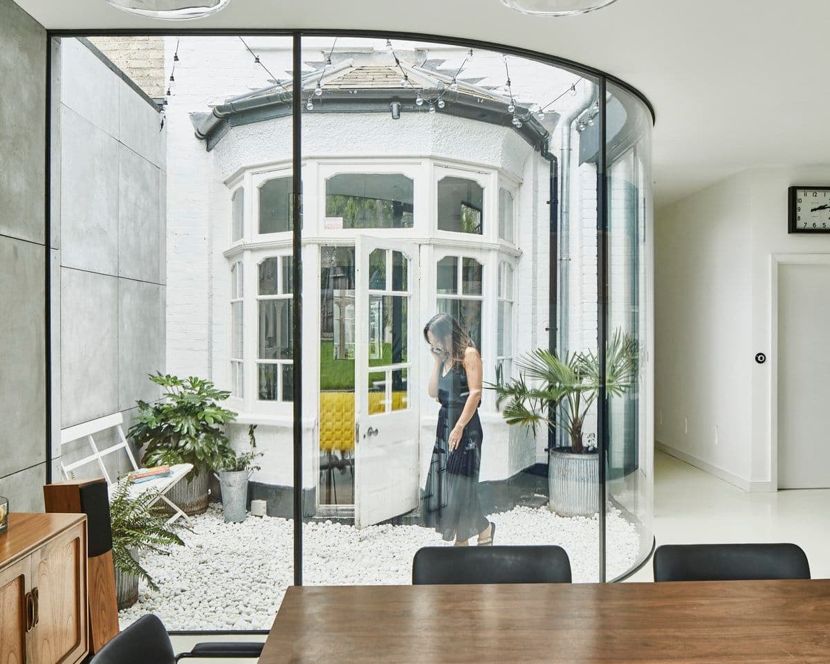 Modern dining area with glass wall overlooking a quaint courtyard with plants and a woman outside.