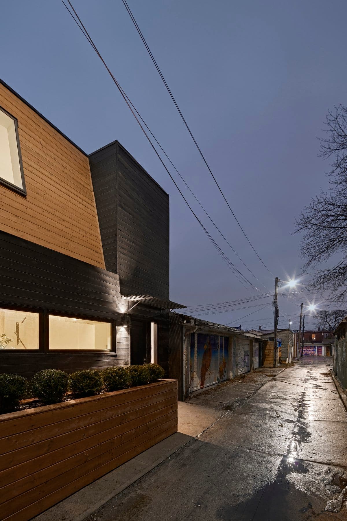 Modern minimalist house in wet alleyway at dusk with warm window light.