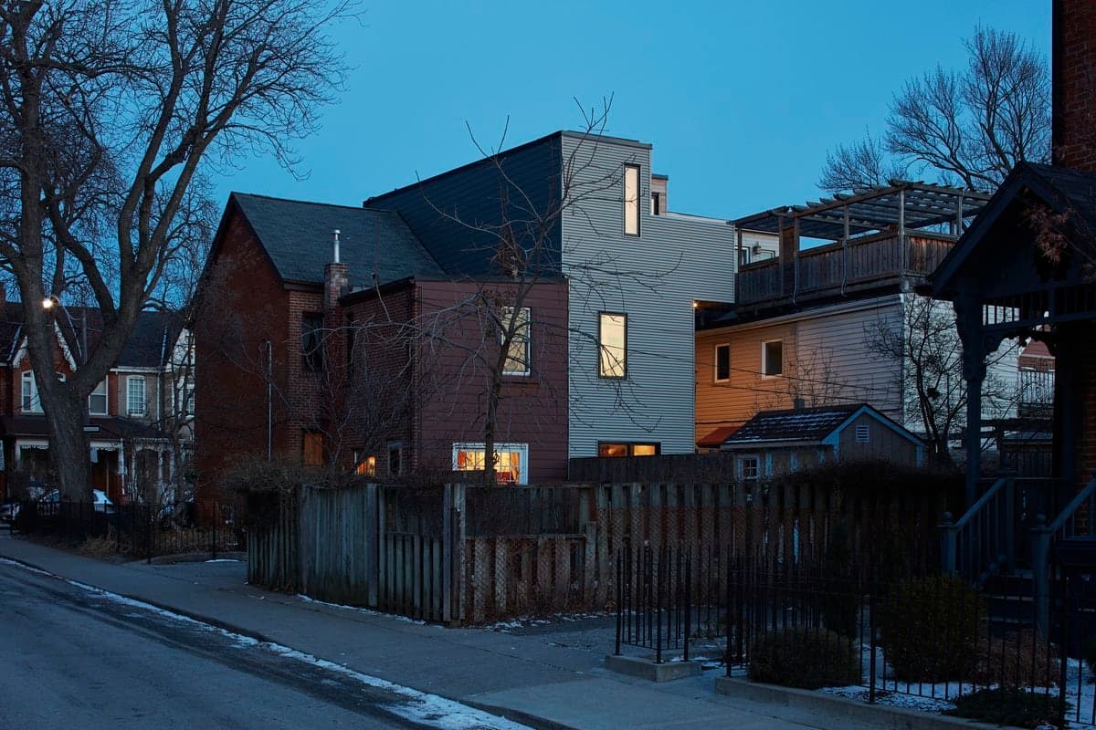 Modern three-story house at dusk with older brick house adjacent in urban residential street.