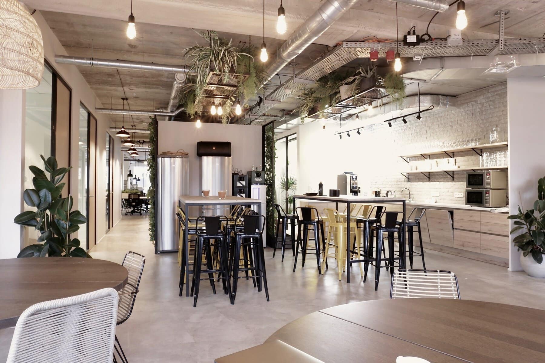 Modern industrial office kitchen with open-concept design, wooden tables, bar stools, and stainless steel appliances.