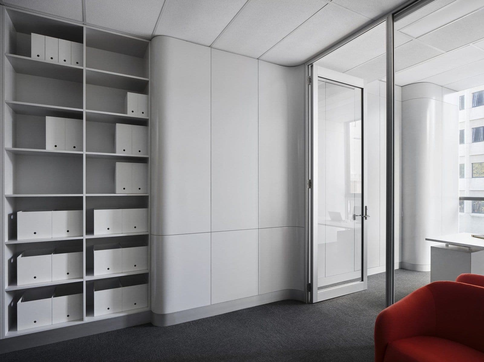 Modern minimalistic office with white storage, glass door, red armchair, and dark flooring.