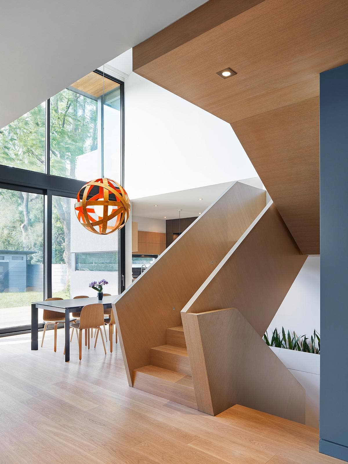 Modern minimalist interior with light wood staircase and dining area, featuring bold orange light fixture.