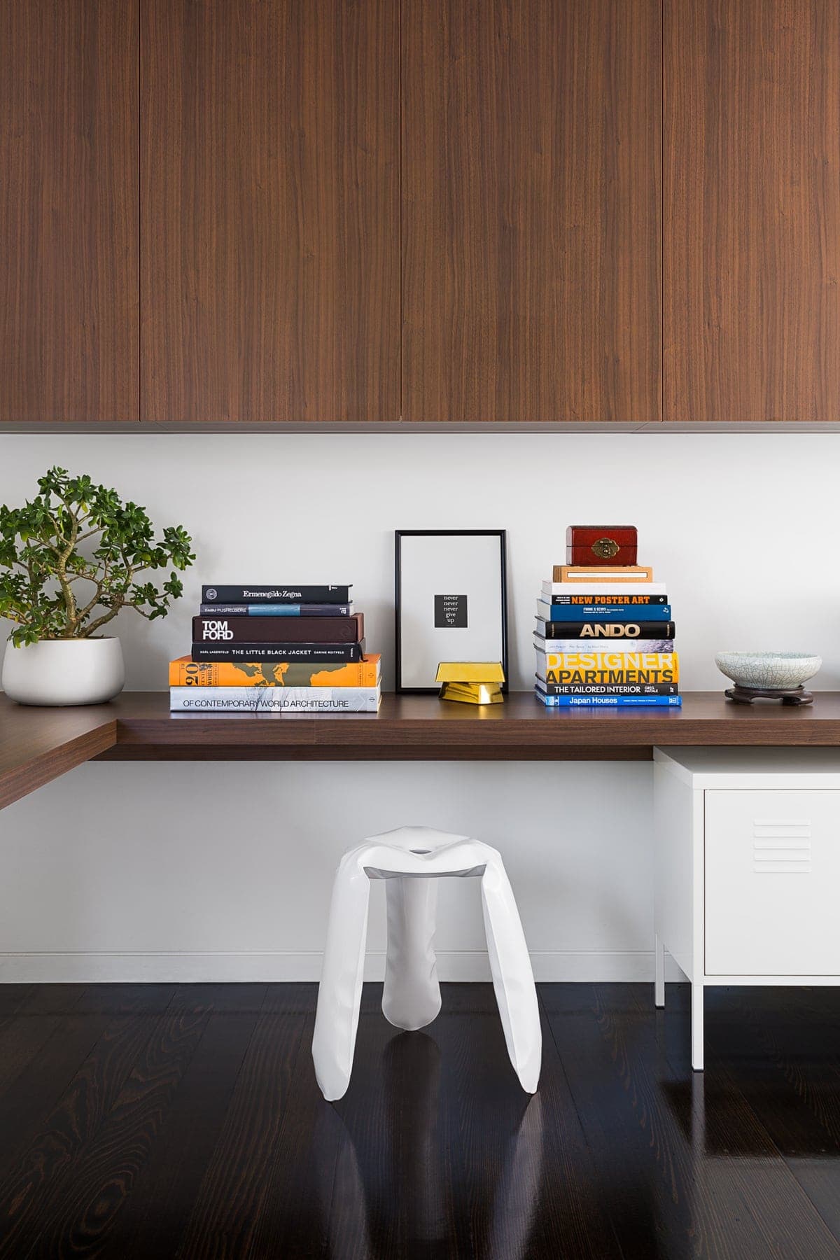 Modern minimalist workspace with wooden desk, potted plant, books, artwork, and stylish white stool.
