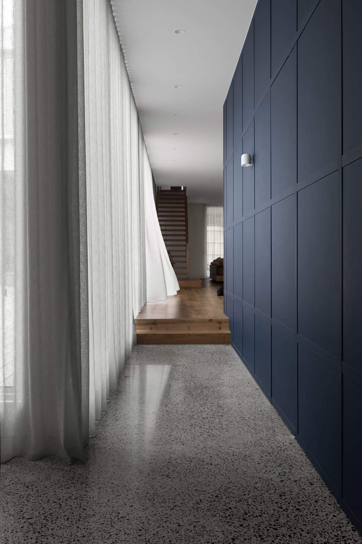 Modern minimalist hallway with polished concrete floor, sheer curtains, blue wall, and wooden staircase.