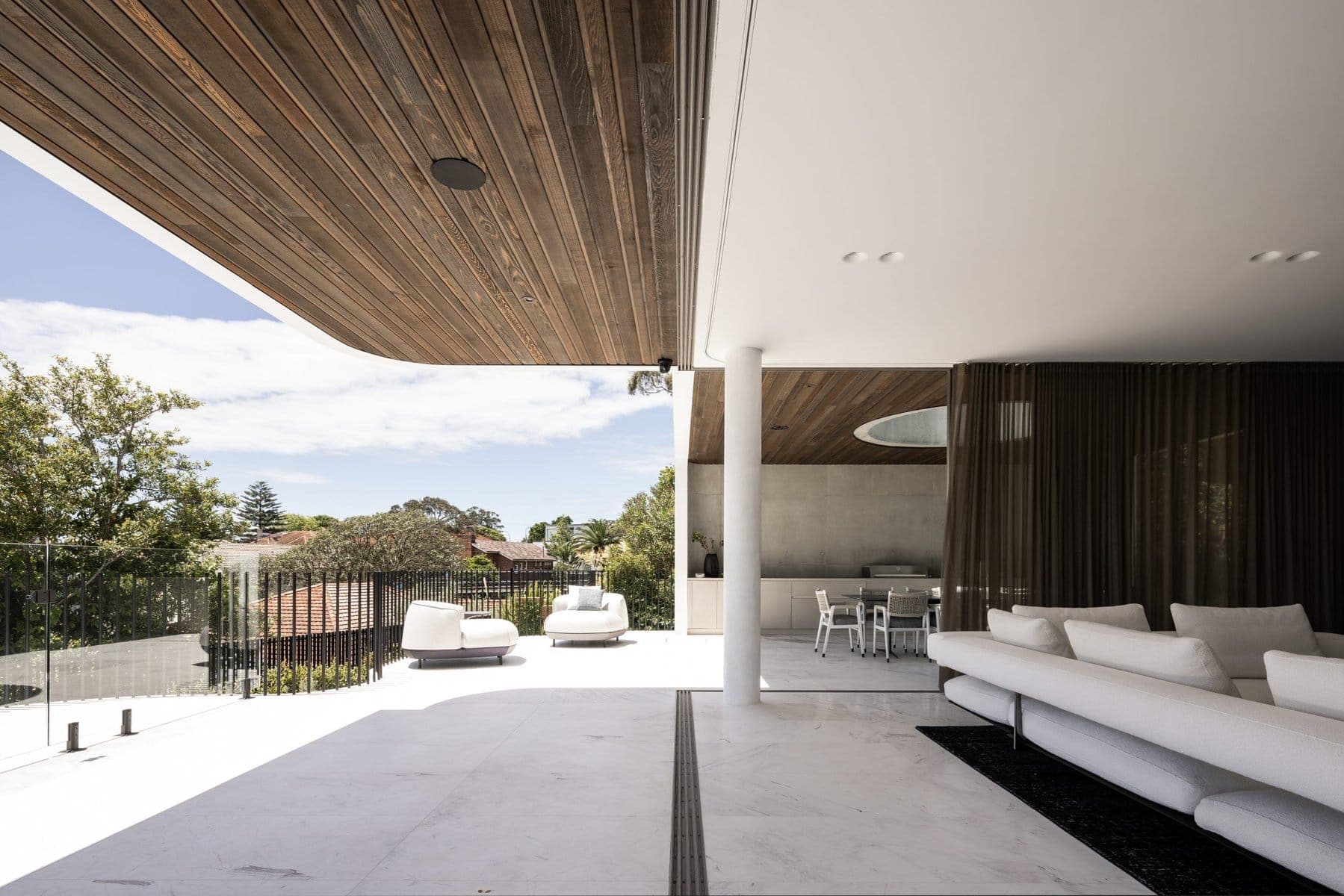 Luxurious modern indoor-outdoor living space with wooden and white ceilings, sleek furniture, and greenery.