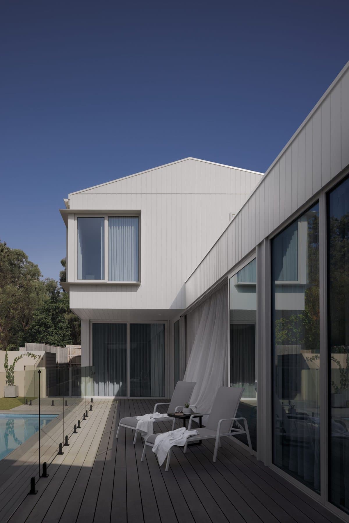 Modern two-level house with minimalist design, large windows, wooden deck, glass railing, and swimming pool.