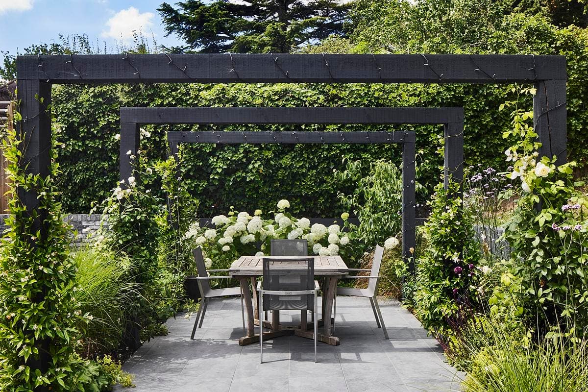 Elegant outdoor dining area in a lush garden with modern furniture and rustic wooden arbors.