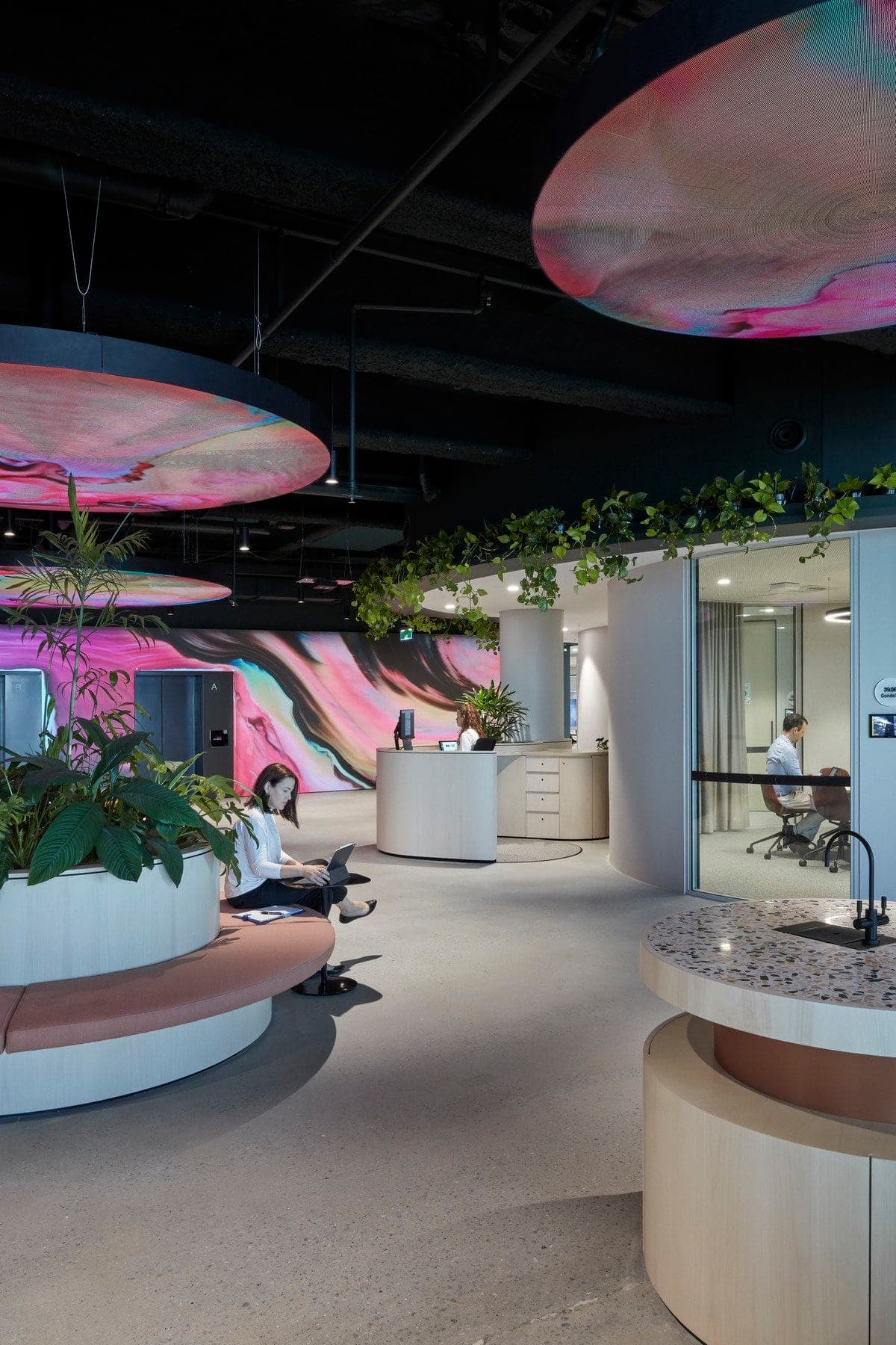 Modern industrial office with plants, circular seating, colorful mural, and glass-walled meeting rooms.