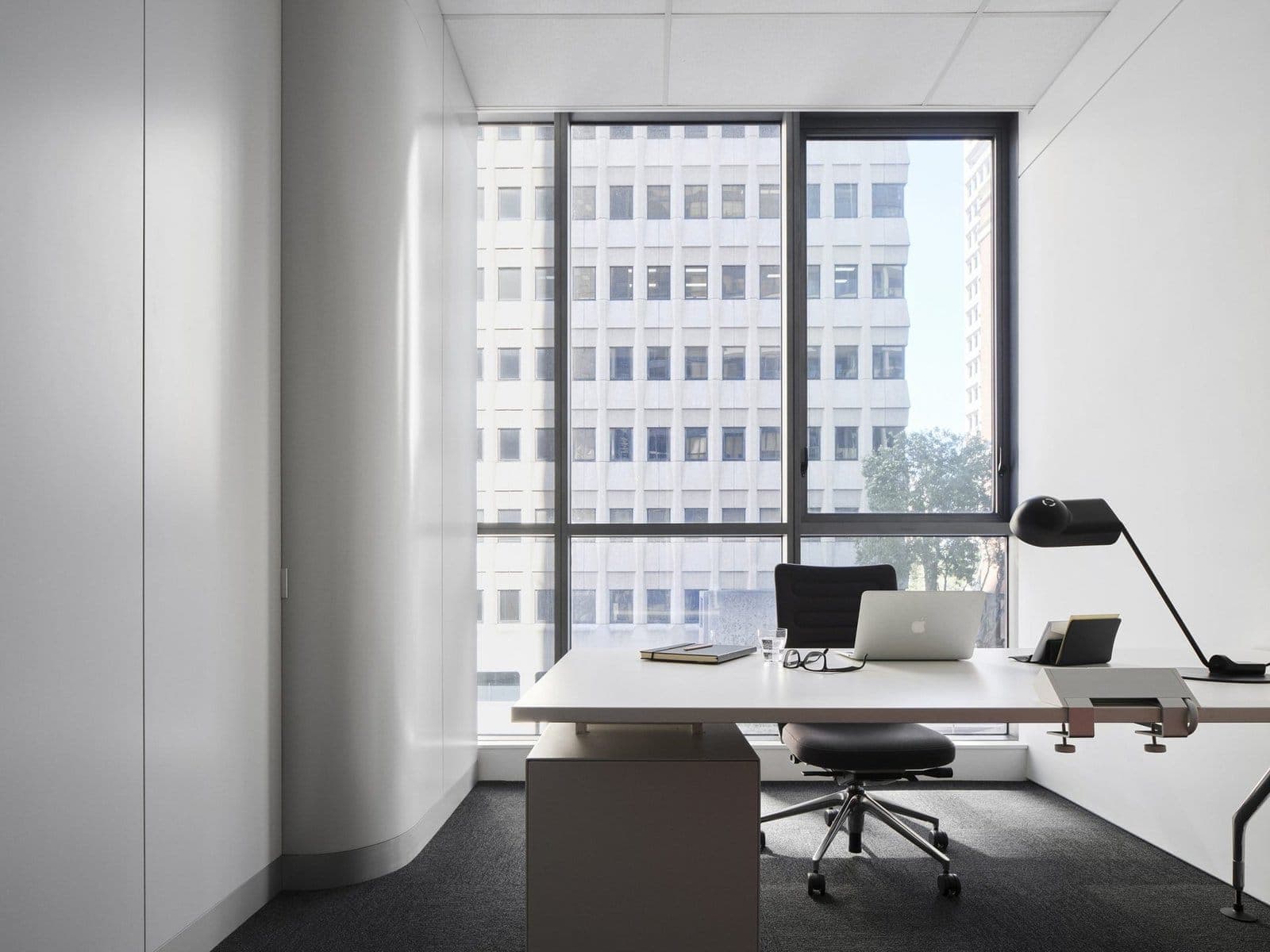 Modern minimalist office with sleek white desk, laptop, and natural light from large window.