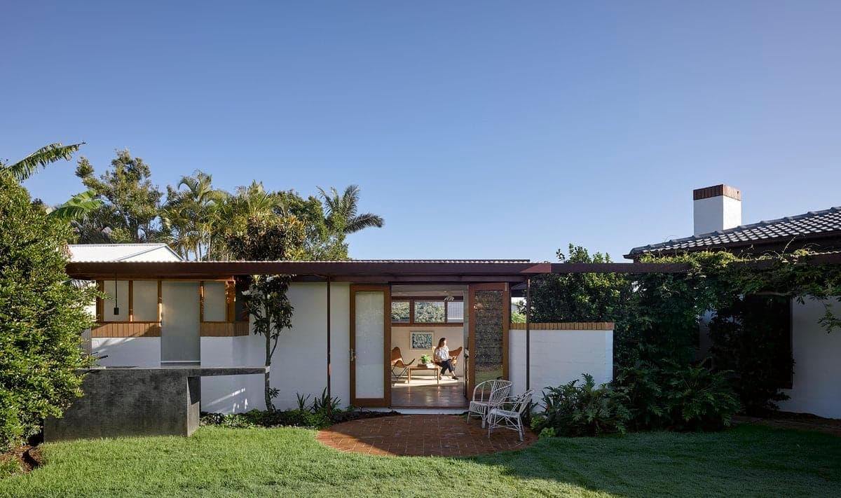 Modern single-story house with white walls, flat roof, wood accents, glass entrance, and lush landscaping.