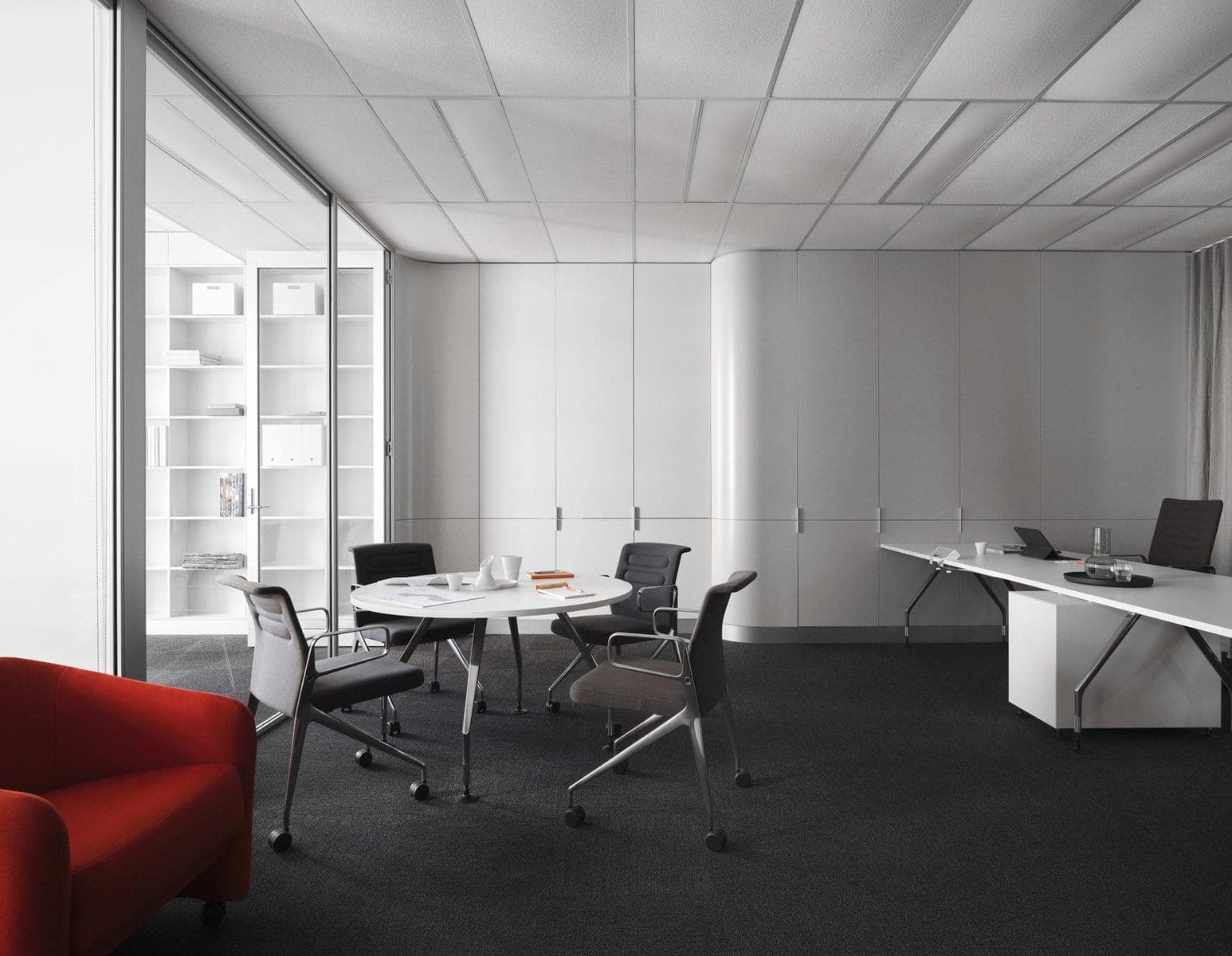 Modern minimalist office with a white desk, meeting table, glass partition, and red armchair.