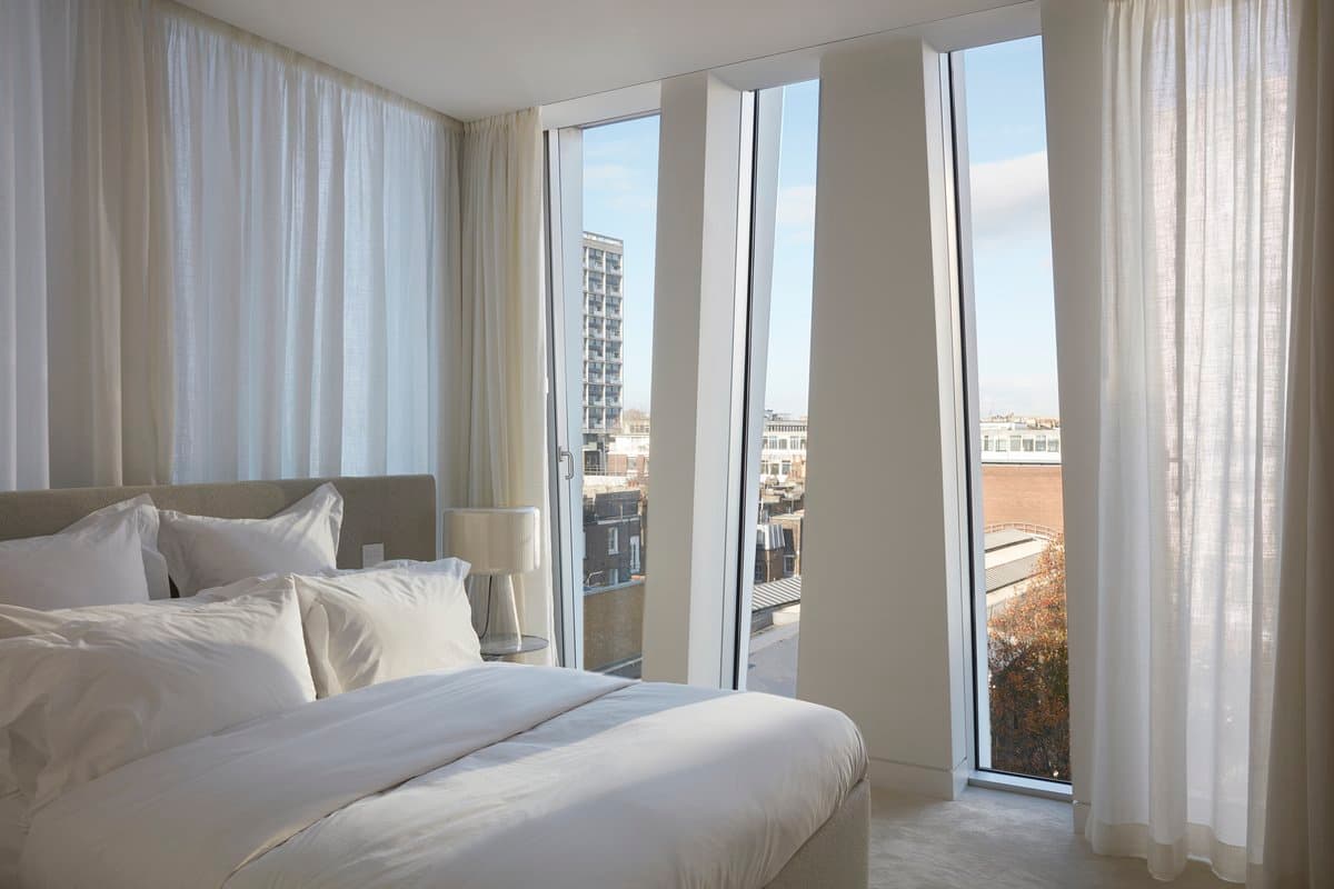 Modern minimalist bedroom with city views, natural light, white linens, and floor-to-ceiling windows.