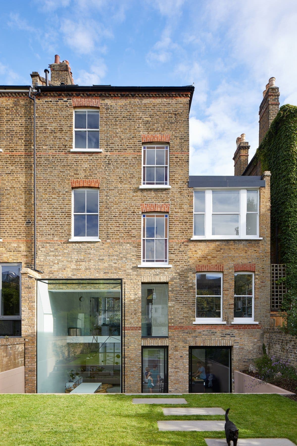 Urban multi-story brick house with modern glass extension, lush garden, and a black cat.
