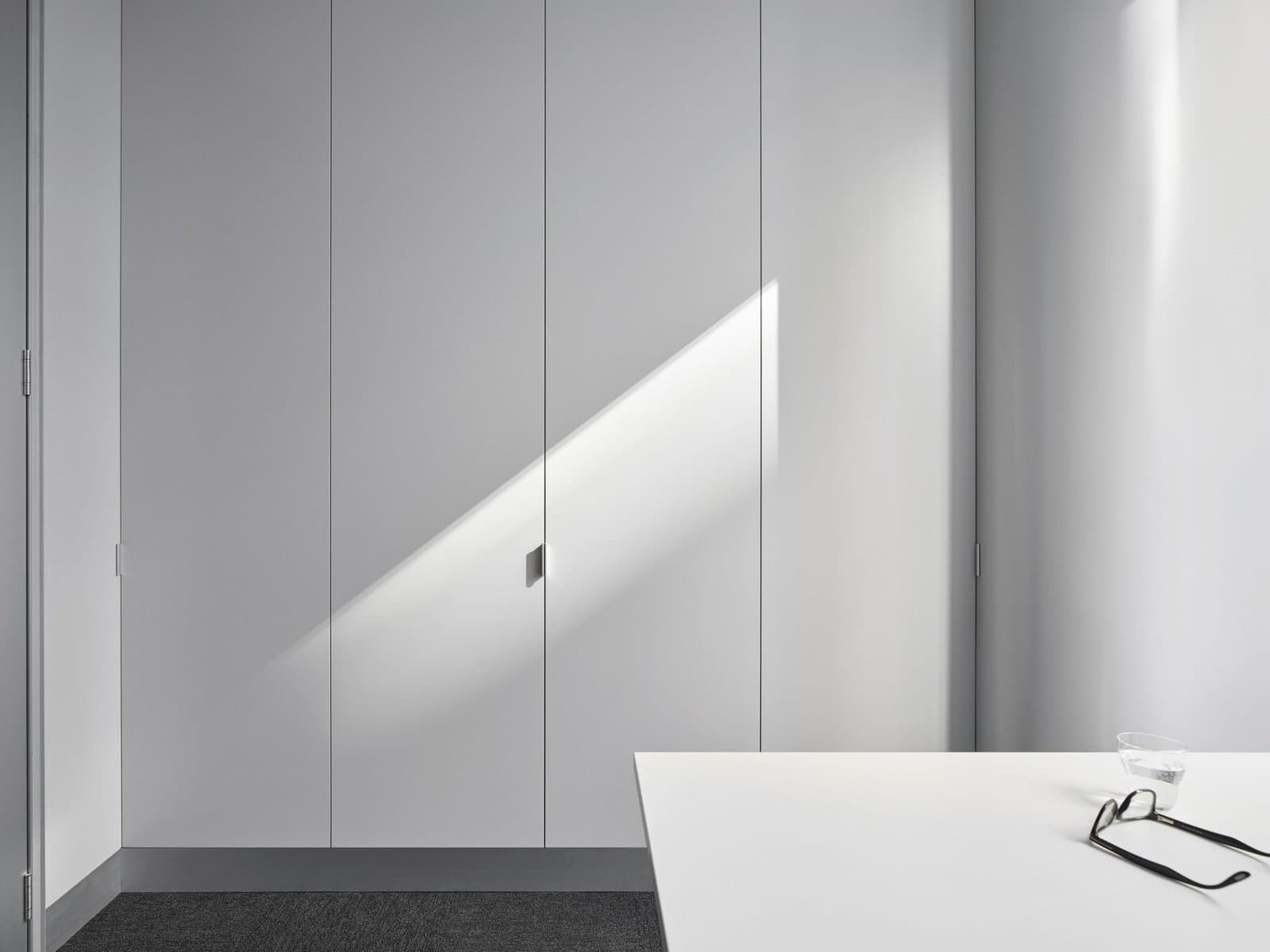 Modern minimalist white office with built-in closet and desk, sunlight creating dynamic lighting.