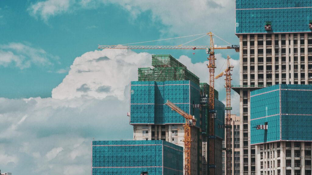 Urban high-rise construction with cranes and blue netting under a partly cloudy sky.