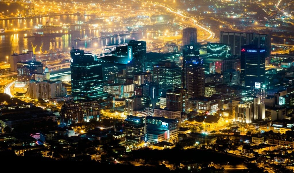 Vibrant night cityscape with illuminated skyscrapers, busy streets, and reflective water body.