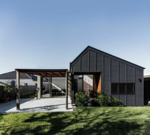 Modern minimalist house with dark cladding, large windows, and shaded outdoor area.