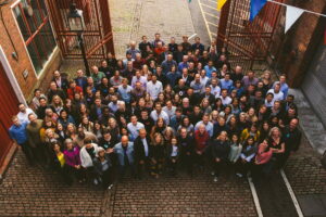 Large diverse group at festive outdoor event in cobblestone courtyard between red brick buildings.