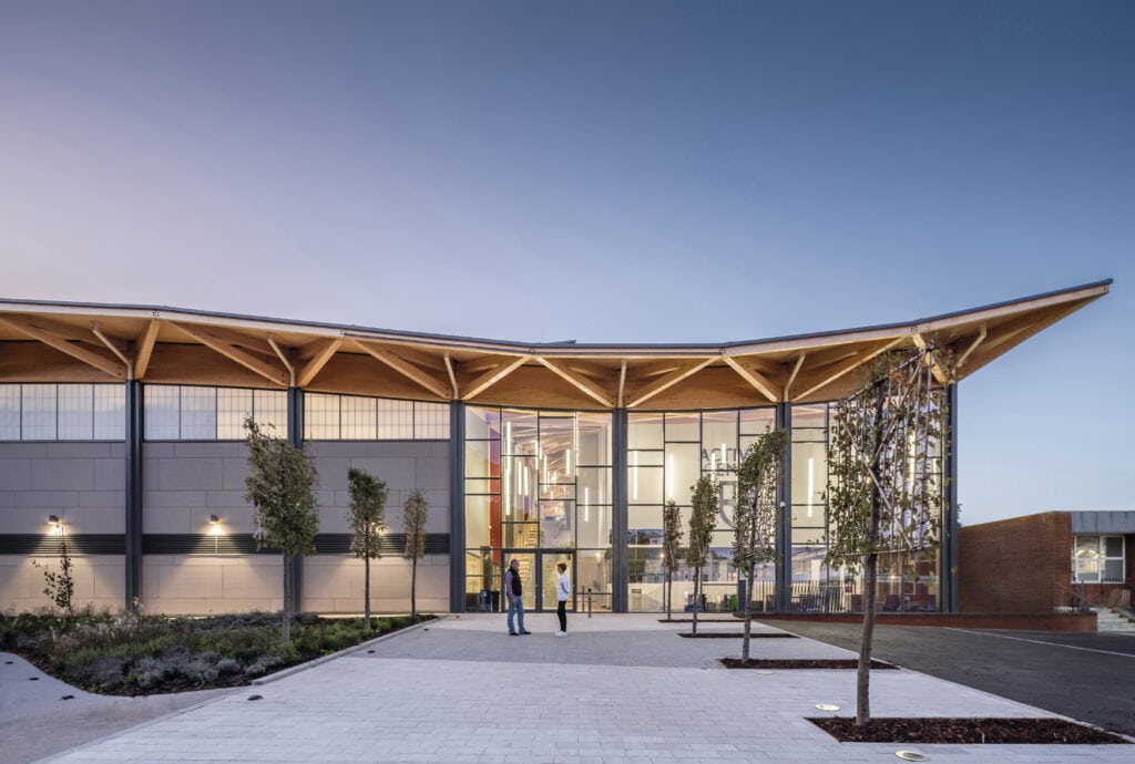 Modern building with sweeping roofline, glass facade, open plaza, and trees; people at entrance.
