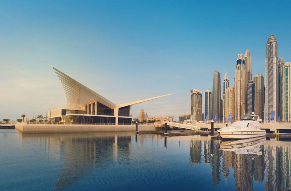 Futuristic waterfront building with yachts, high-rise skyline, and tranquil water reflection.