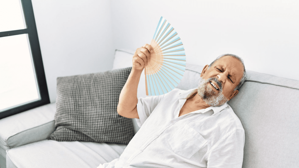 Elderly man fanning himself, struggling with heat in a bright, minimalist living room.