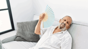 Elderly man fanning himself, struggling with heat in a bright, minimalist living room.