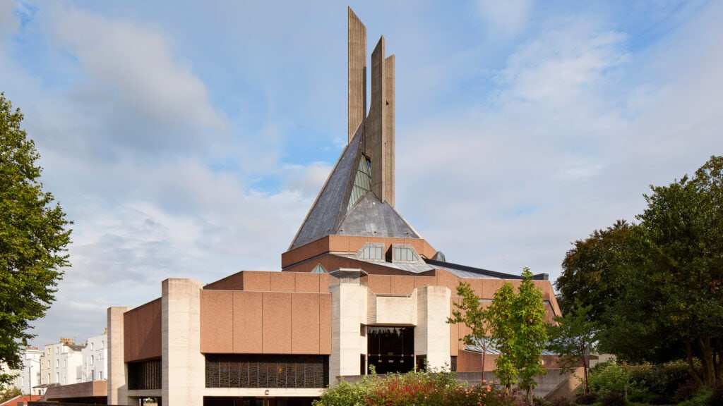 Modern church with angular design, red brick base, reflective roof, and tall spires amidst urban greenery.