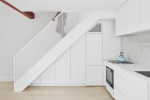 Modern white kitchen under stairs with glass railing and light wood flooring.