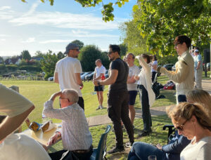Group enjoying sunny day in lush park, engaging in casual activities, shaded by trees.