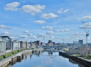 Modern urban skyline with river, bridge, and diverse buildings reflecting blue sky.