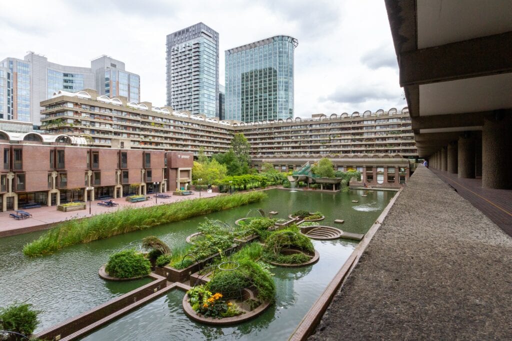 Modern urban landscape with pond, greenery, and contemporary buildings reflecting in clear water.