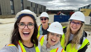 Four women smiling at a construction site, wearing hard hats and safety vests.